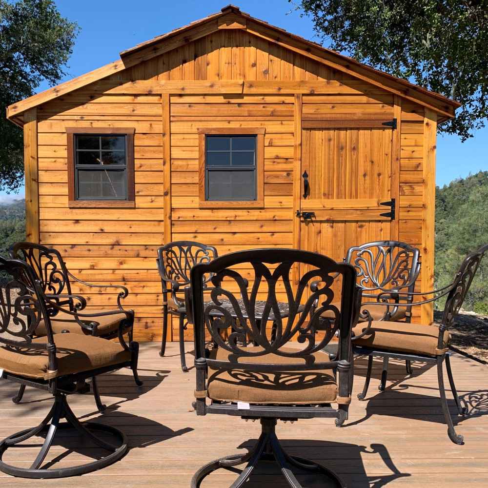 An Outdoor Living Today 12×12 Sunshed Garden Shed stands behind a set of elegant patio chairs on a deck