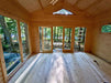 An empty wood interior of the Sawmill Structures Edwin Lake House 70MM Garden Room shows a peaceful view through full glass doors out to a wooded area