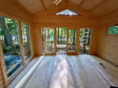 An empty wood interior of the Sawmill Structures Edwin Lake House 70MM Garden Room shows a peaceful view through full glass doors out to a wooded area