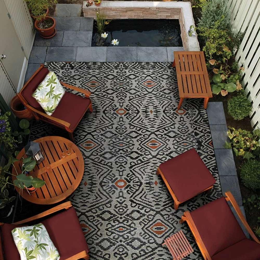An outdoor patio scene features Couristan Dolce Indoor/Outdoor Rug - Mala/Smoke beneath wooden furniture and near a koi pond
