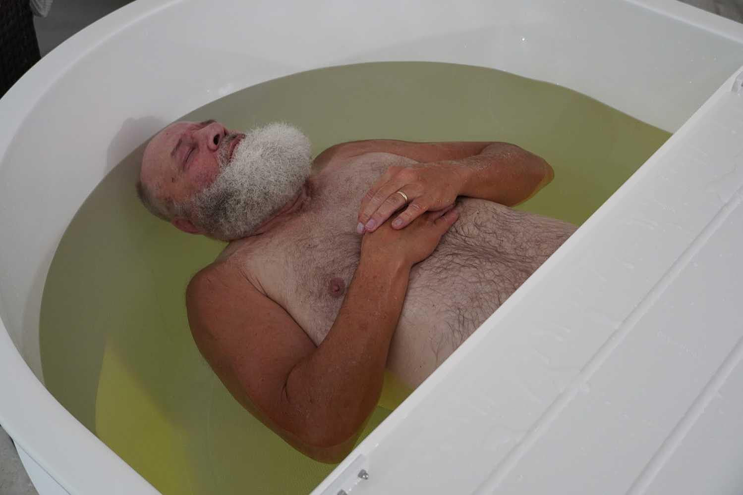 An elderly man with a white beard lies peacefully in a Dreampod Home Float Pro filled with water, appearing deeply relaxed