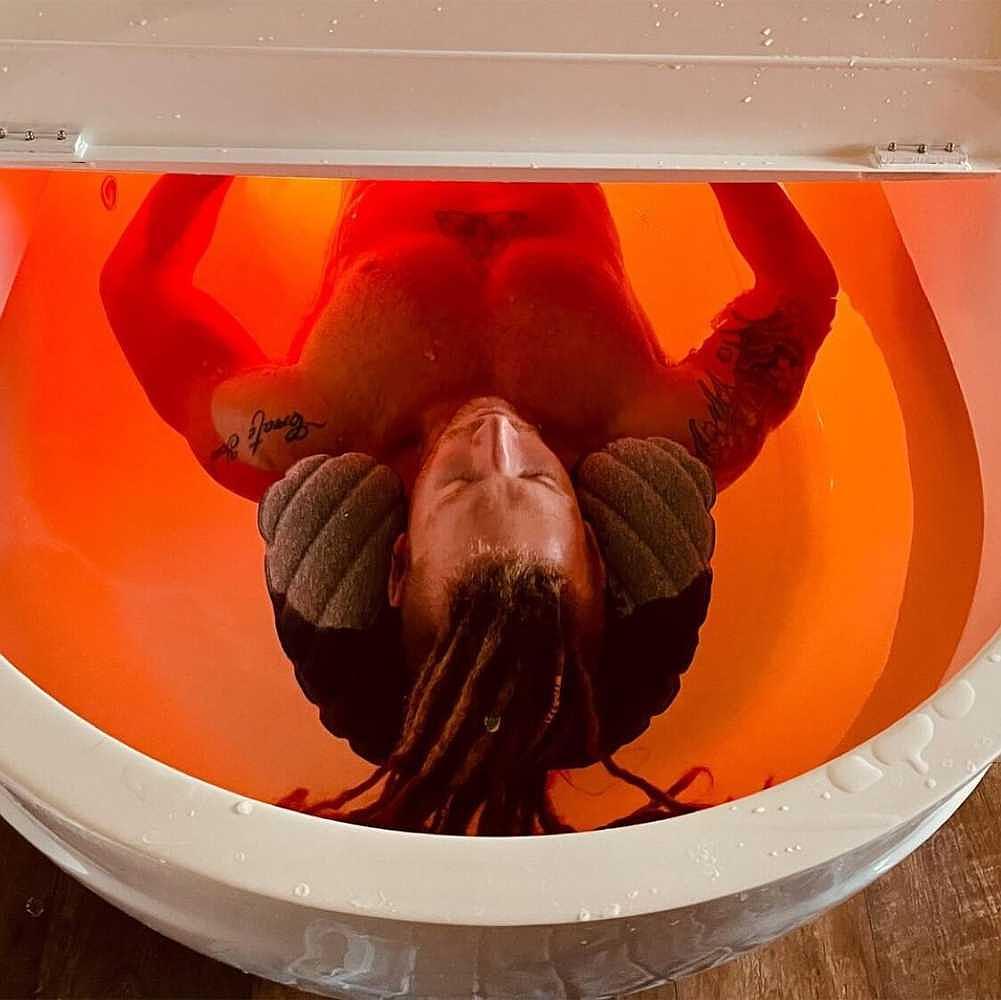 A person with dreadlocks floats serenely in a Dreampod Home Float Pro, with the water illuminated in a warm red glow