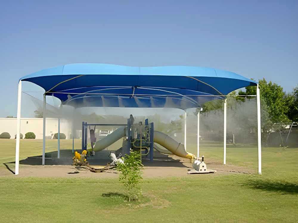A playground under a large blue canopy is cooled by the Aeromist Pinnacle 1000 PSI Misting System releasing mist above the play structure