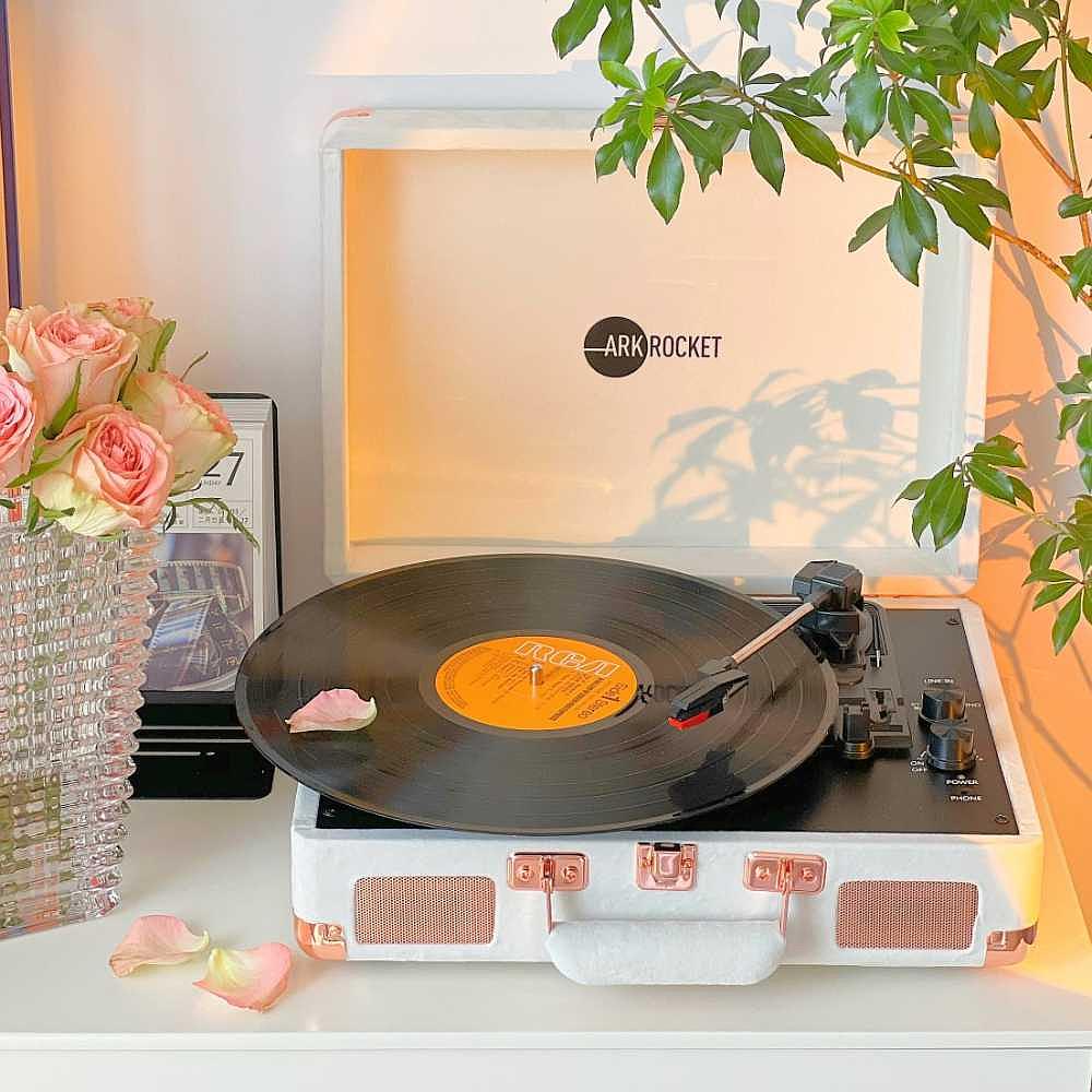 Arkrocket Curiosity Suitcase Bluetooth Turntable (Cream Velvet) is elegantly displayed on a table next to pink roses