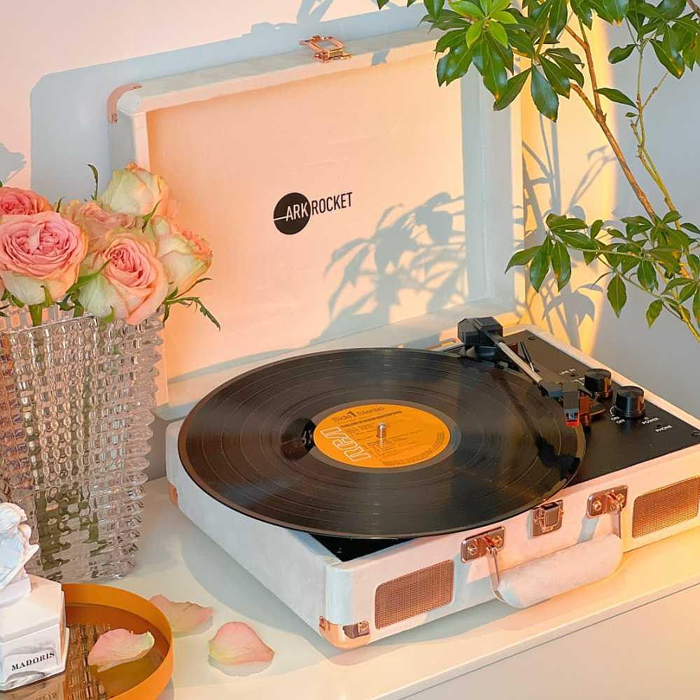 Arkrocket Curiosity Suitcase Bluetooth Turntable (Cream Velvet) is set on a table surrounded by roses playing a record