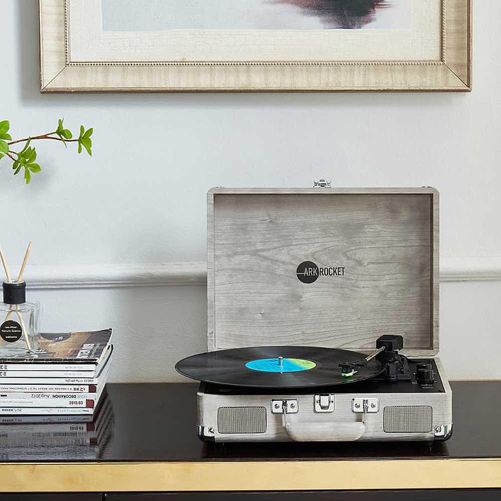 Arkrocket Curiosity Suitcase Bluetooth Turntable (Gray Wood Grain) sits open on a sleek wooden cabinet with a vinyl record spinning