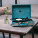 Arkrocket Curiosity Suitcase Bluetooth Turntable (Green Velvet) is open on a table playing a record with flowers in a vase nearby