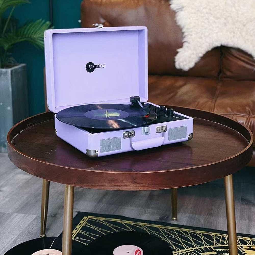 Arkrocket Curiosity Suitcase Bluetooth Turntable (Lavender) is placed on a round wooden table, playing a vinyl record in a cozy room setting