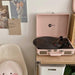 Arkrocket Curiosity Suitcase Bluetooth Turntable (Pale Pink) is set up on a small wooden table with a vinyl spinning and decor on the wall behind it