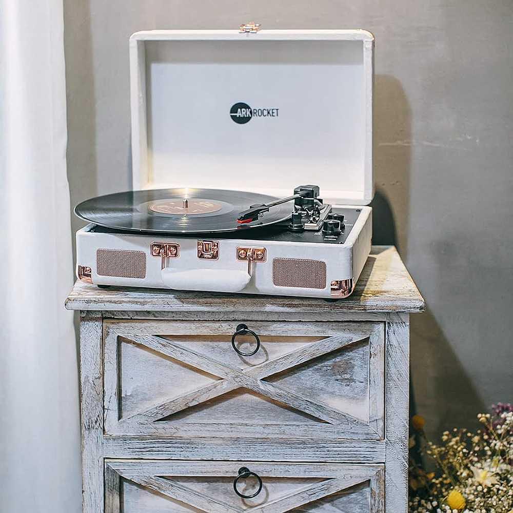 Arkrocket Curiosity Suitcase Bluetooth Turntable (White) is sitting on a rustic wooden cabinet playing a record with its lid open