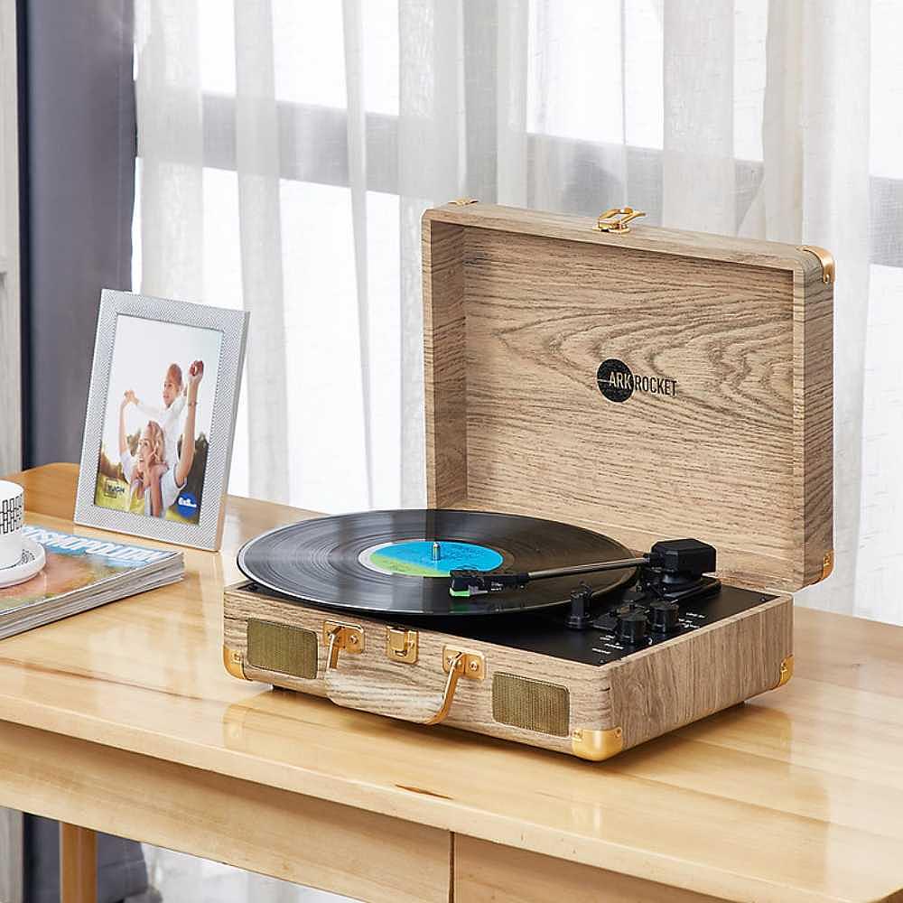 Arkrocket Curiosity Suitcase Bluetooth Turntable (Wood Grain) is placed on a table while a vinyl record spins