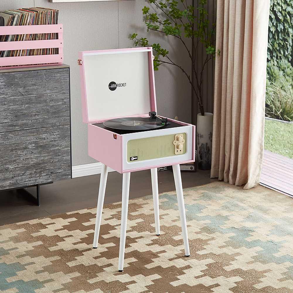 Arkrocket Discovery Bluetooth Record Player Stand with 3-Speed Turntable (PinkWhite) is displayed in a modern room on tall white legs beside a window and record holder