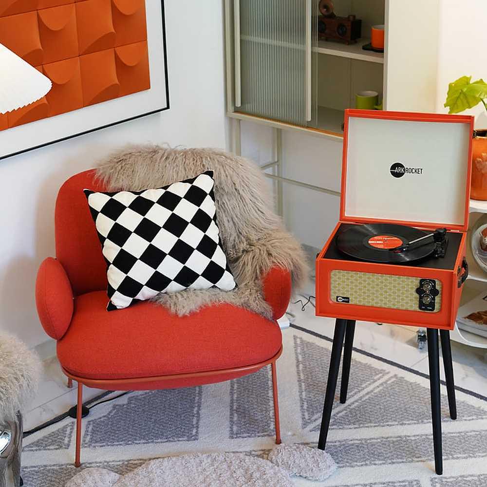 Arkrocket Discovery II Bluetooth Turntable Record Player With Legs (Orange) is placed near a cozy red chair