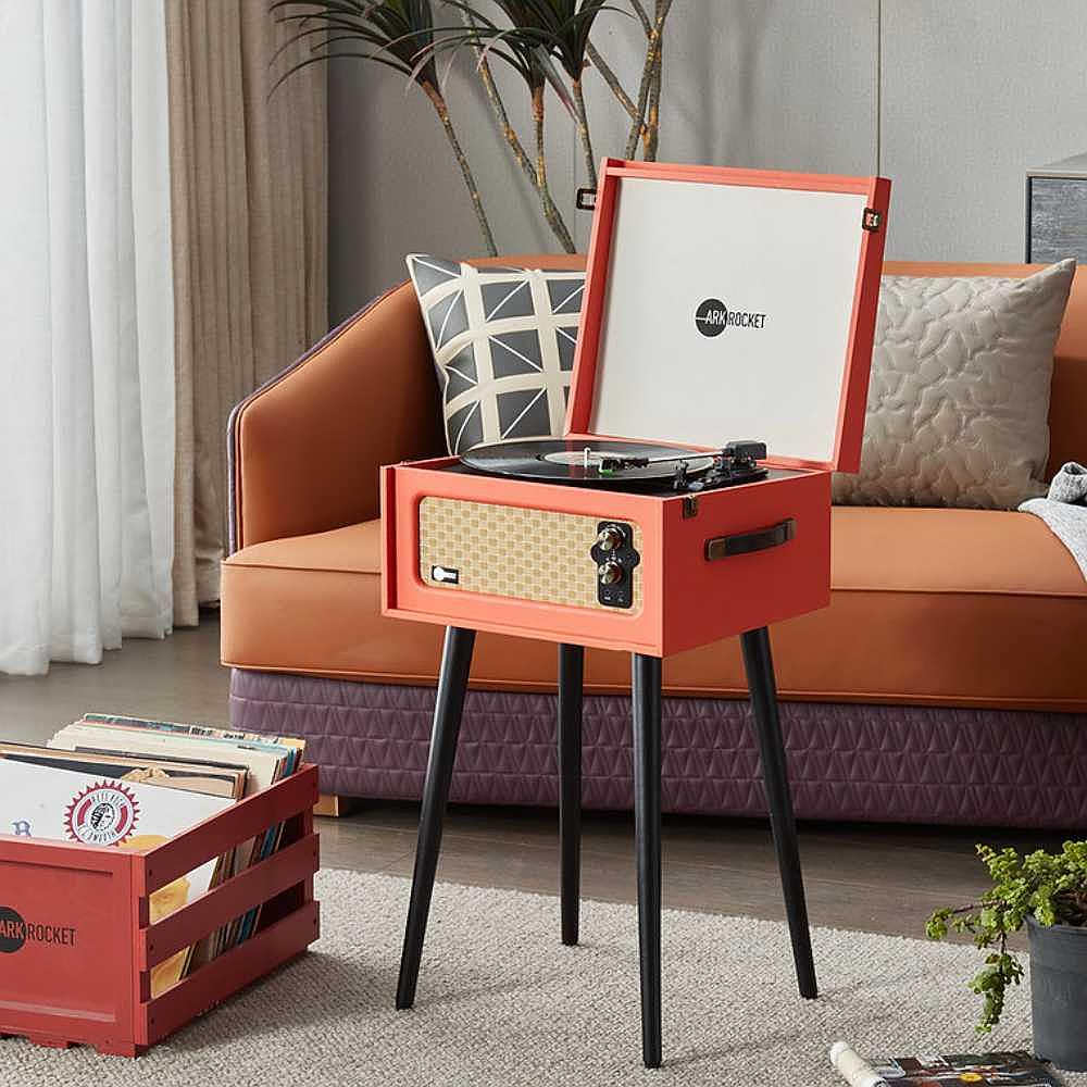 Arkrocket Discovery II Bluetooth Turntable Record Player With Legs (Orange) sits stylishly next to an orange couch in a modern living room