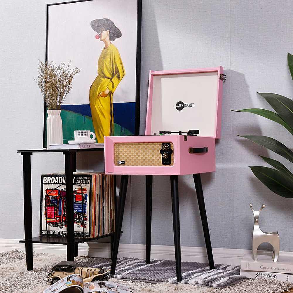 Arkrocket Discovery II Bluetooth Turntable Record Player With Legs (Pink) is displayed in a room next to a framed painting and a small table with records