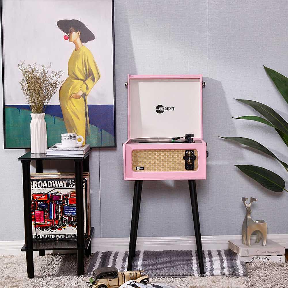 Arkrocket Discovery II Bluetooth Turntable Record Player With Legs (Pink) is displayed in a room setting with a painting on the wall and decorative items