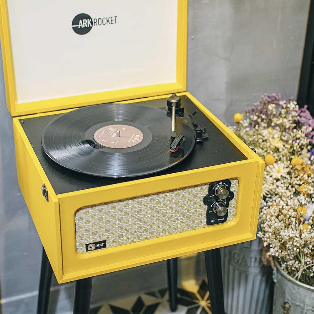 Arkrocket Retro Turntable Wireless Record Player With Legs (Yellow) is positioned next to a vase of flowers with a vinyl record playing