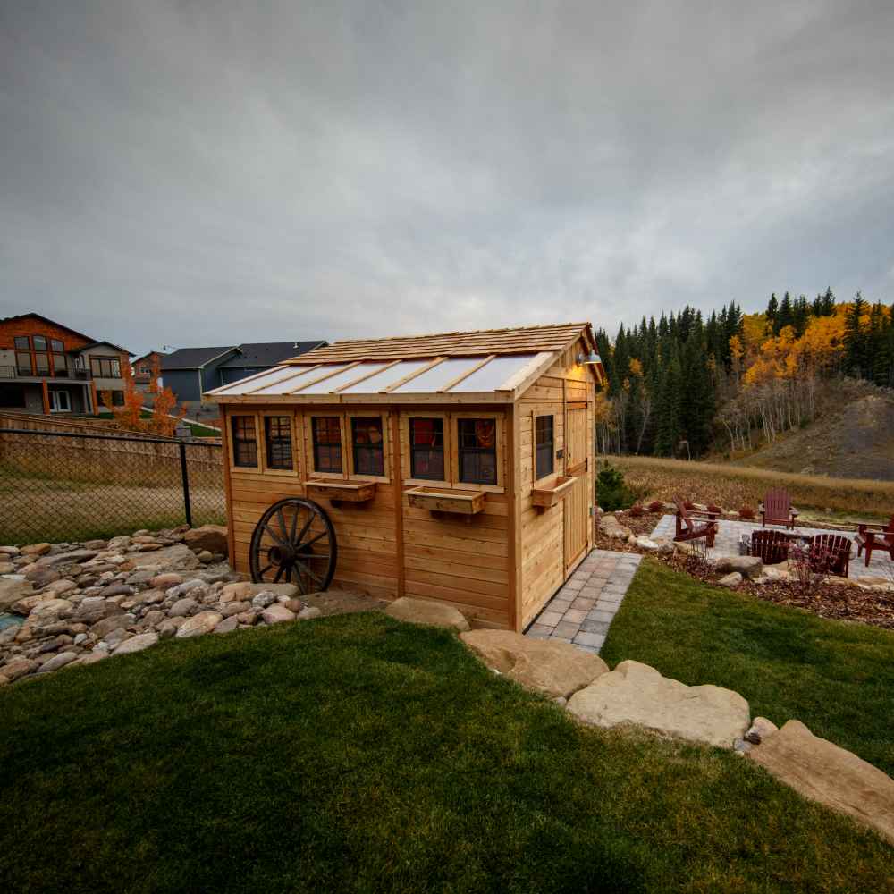 A rustic backyard scene shows the Outdoor Living Today 8×12 Sunshed Garden Shed with a cedar finish, wagon wheel accent, and fire pit seating nearby under a cloudy sky