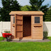 A rustic garden shed with one open door, cedar roof, and a red wheelbarrow beside it sits on a lawn, representing the Outdoor Living Today 9×6 Cabana Garden Shed