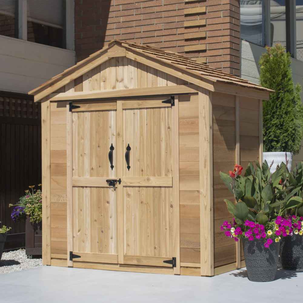 A side-angled view of a closed Outdoor Living Today SpaceMaster 6×4 with Double Door displays its natural wood exterior and black metal door handles next to flower pots