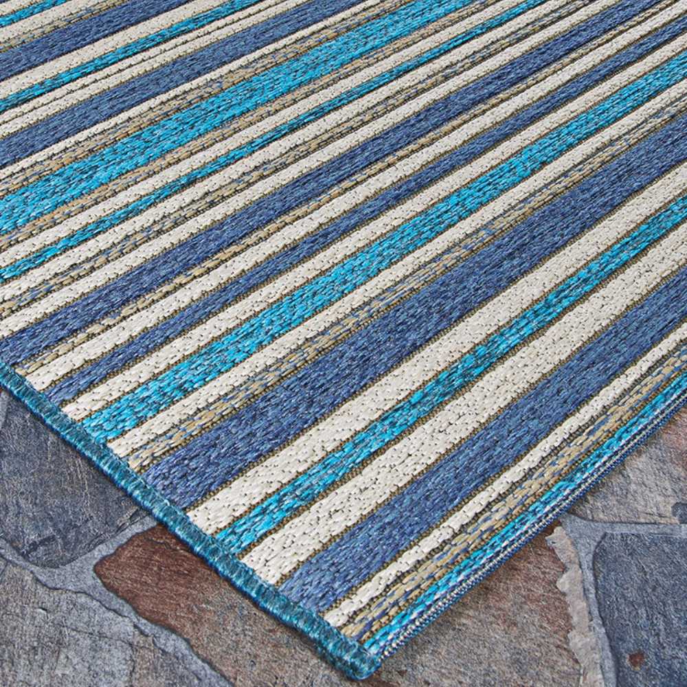 A side angle of the Couristan Cape Indoor/Outdoor Rug - Brockton/Cobalt-Teal displays its vivid linear design with various shades of blue and beige on a stone surface