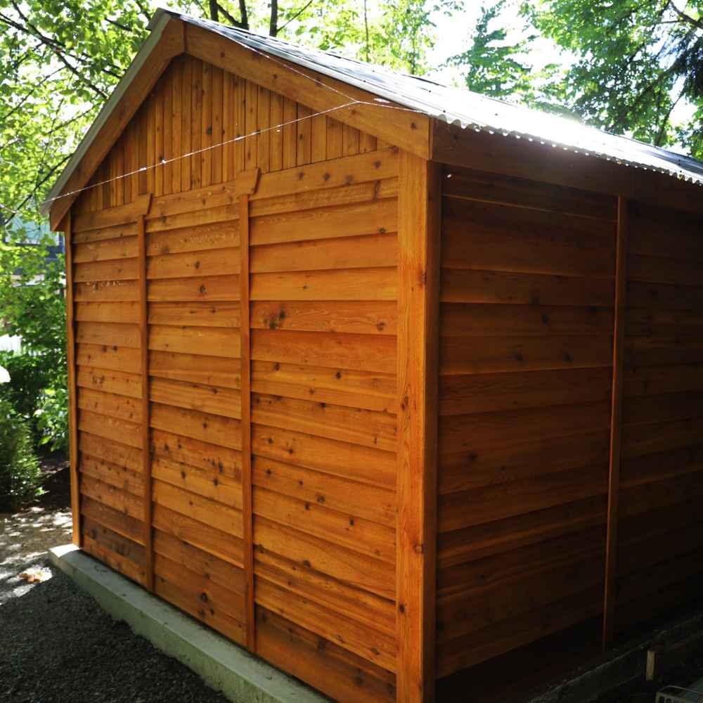 A side angle view of the Outdoor Living Today 12×12 Double Door Space Master Storage Shed shows its beautifully crafted cedar wood panels under a sloped black roof