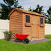 A side view of the Outdoor Living Today 9×6 Cabana Garden Shed features twin windows and cedar roof with flower boxes and a red wheelbarrow on a grassy yard