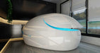 A sleek, white Dreampod Vmax Float Pod with a futuristic design and a glowing blue accent, shown partially open in a modern room