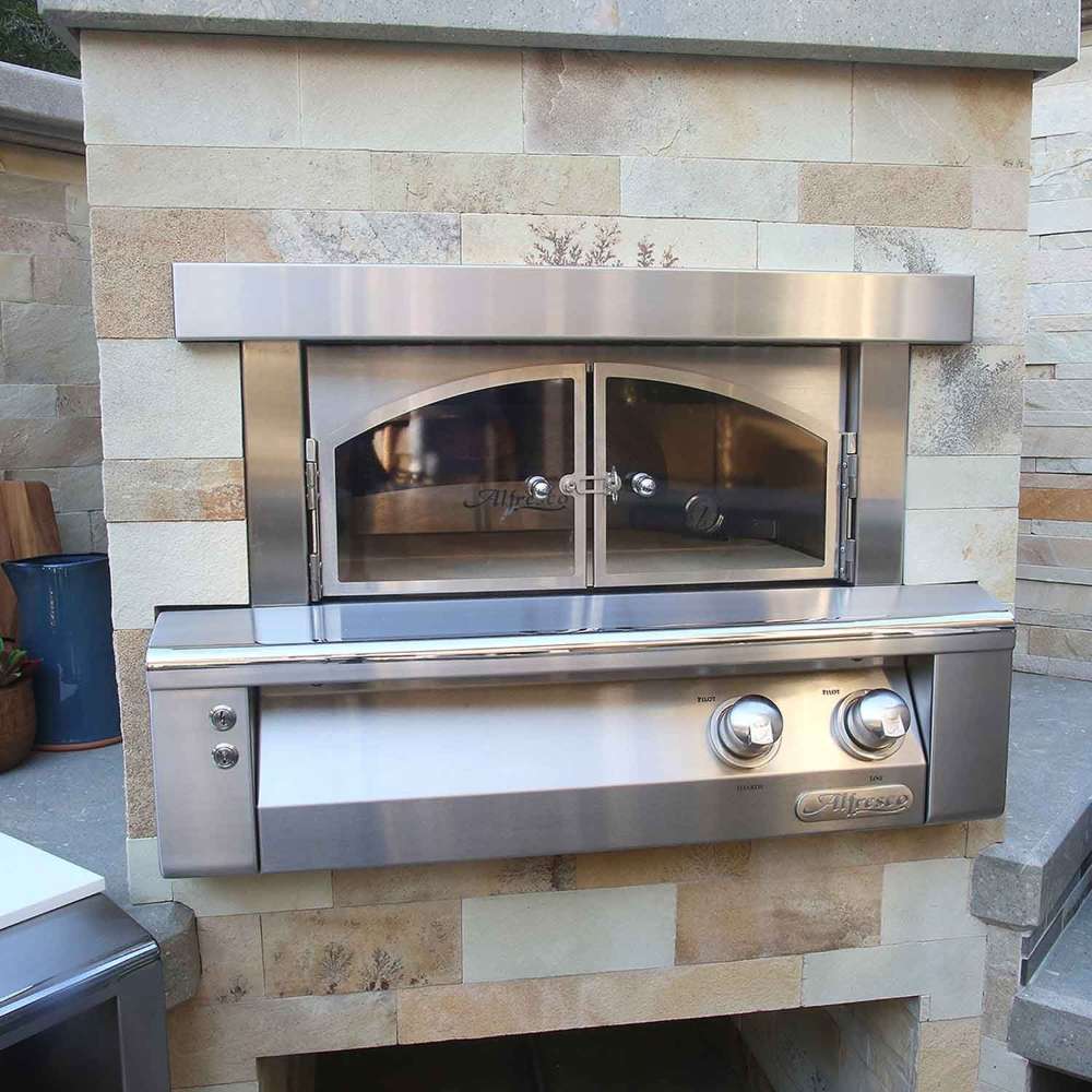 A straight-on view of the Alfresco Grills 30 Pizza Oven Plus - Built-In displays its stainless steel construction and arched glass doors against a stone wall