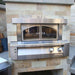 A straight-on view of the Alfresco Grills 30 Pizza Oven Plus - Built-In displays its stainless steel construction and arched glass doors against a stone wall