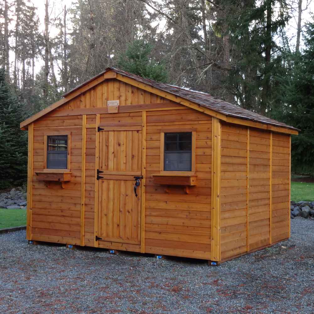 A sturdy brown Outdoor Living Today 12×12 Sunshed Garden Shed with a simple gable roof and two front-facing windows sits on a gravel pad surrounded by tall trees