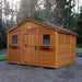 A sturdy brown Outdoor Living Today 12×12 Sunshed Garden Shed with a simple gable roof and two front-facing windows sits on a gravel pad surrounded by tall trees