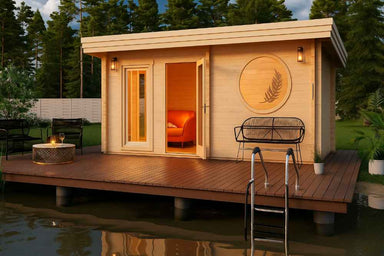 At dusk, the GardenHouse24 USA Sauna Sahib 70 Garden Room glows warmly inside, featuring a round wall engraving and deck seating next to a serene body of water