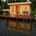 At dusk, the GardenHouse24 USA Sauna Sahib 70 Garden Room glows warmly inside, featuring a round wall engraving and deck seating next to a serene body of water