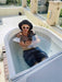 A tattooed man sits comfortably in a Dreampod Ice Bath with Chiller on a balcony with bright sunlight and a view of lush greenery around him