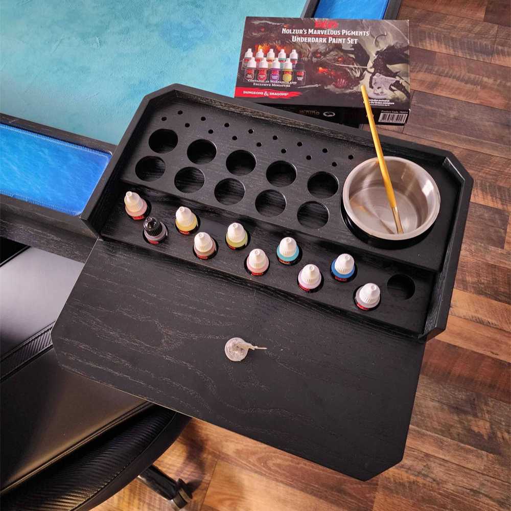 A top-down view of the onyx Game Theory Tables Origins Minature Work Station shows neatly arranged paint slots, a metal brush holder, and room for miniature models