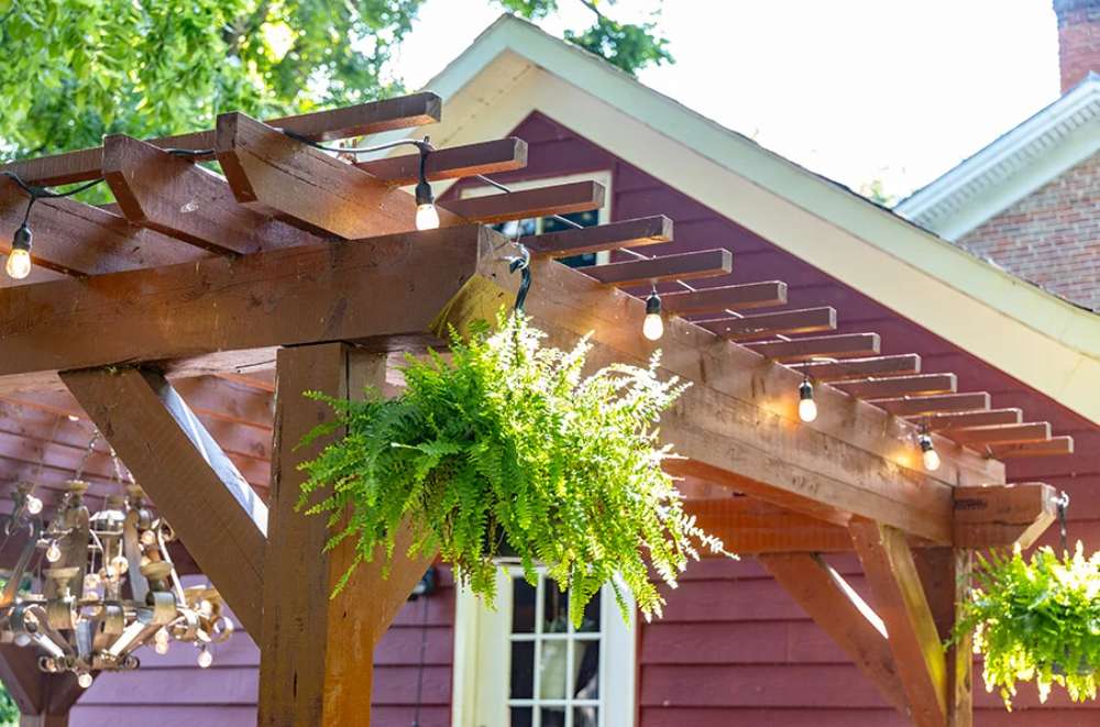 Attached to a red house, the Brookside Timber Frame Shadelan Retreat Pergola features warm string lights and vibrant greenery