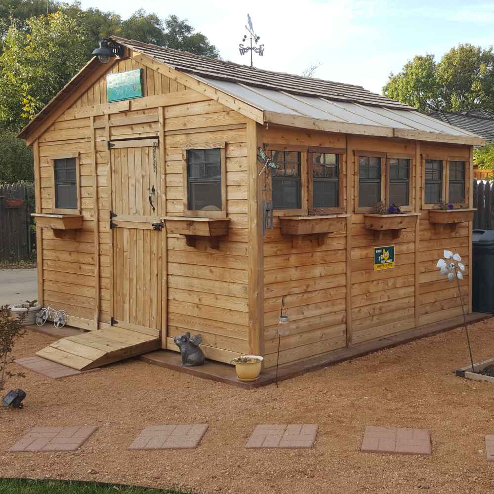 A well-used Outdoor Living Today 12×12 Sunshed Garden Shed with seven small windows and planters along one side stands on reddish gravel with potted plants and yard decor