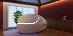 A white Dreampod Vmax Float Pod is illuminated in a warm, wood-paneled room with a plant and soft curtains in the background