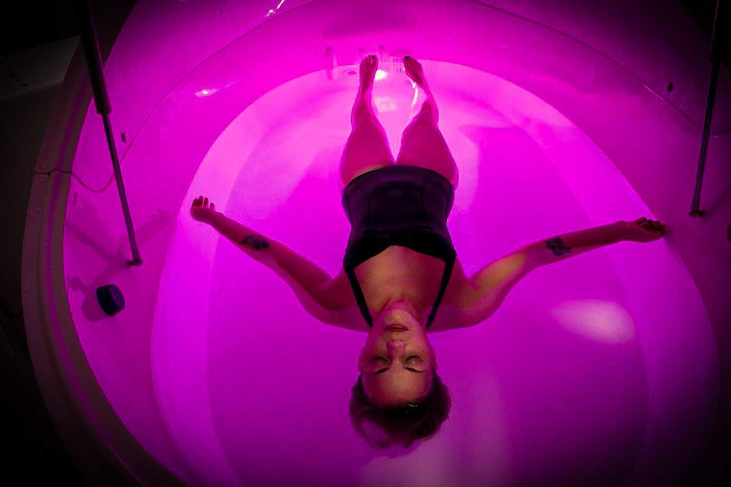 A woman floating in the Dreampod Vmax Float Pod under vibrant pink lighting, with her arms outstretched and relaxed