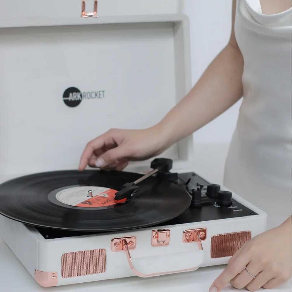 A woman in a white dress is adjusting a record on the Arkrocket Curiosity Suitcase Bluetooth Turntable (Cream Velvet)