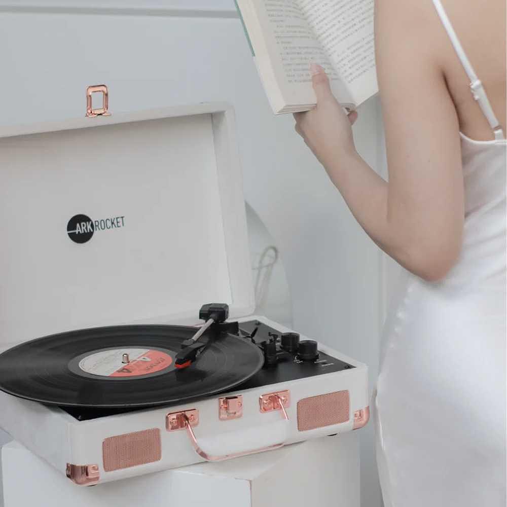 A woman in a white dress is reading a book while a record plays on the Arkrocket Curiosity Suitcase Bluetooth Turntable (Cream Velvet)