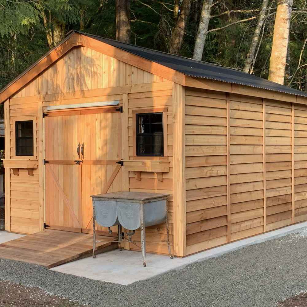 A wooden Outdoor Living Today 12×12 Double Door Space Master Storage Shed stands on a gravel lot with black-framed windows and a weathered metal tub in front