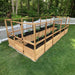 A wooden Outdoor Living Today 8×16 Raised Garden Bed filled with young plants stands on green grass next to a white picket fence in a forested yard