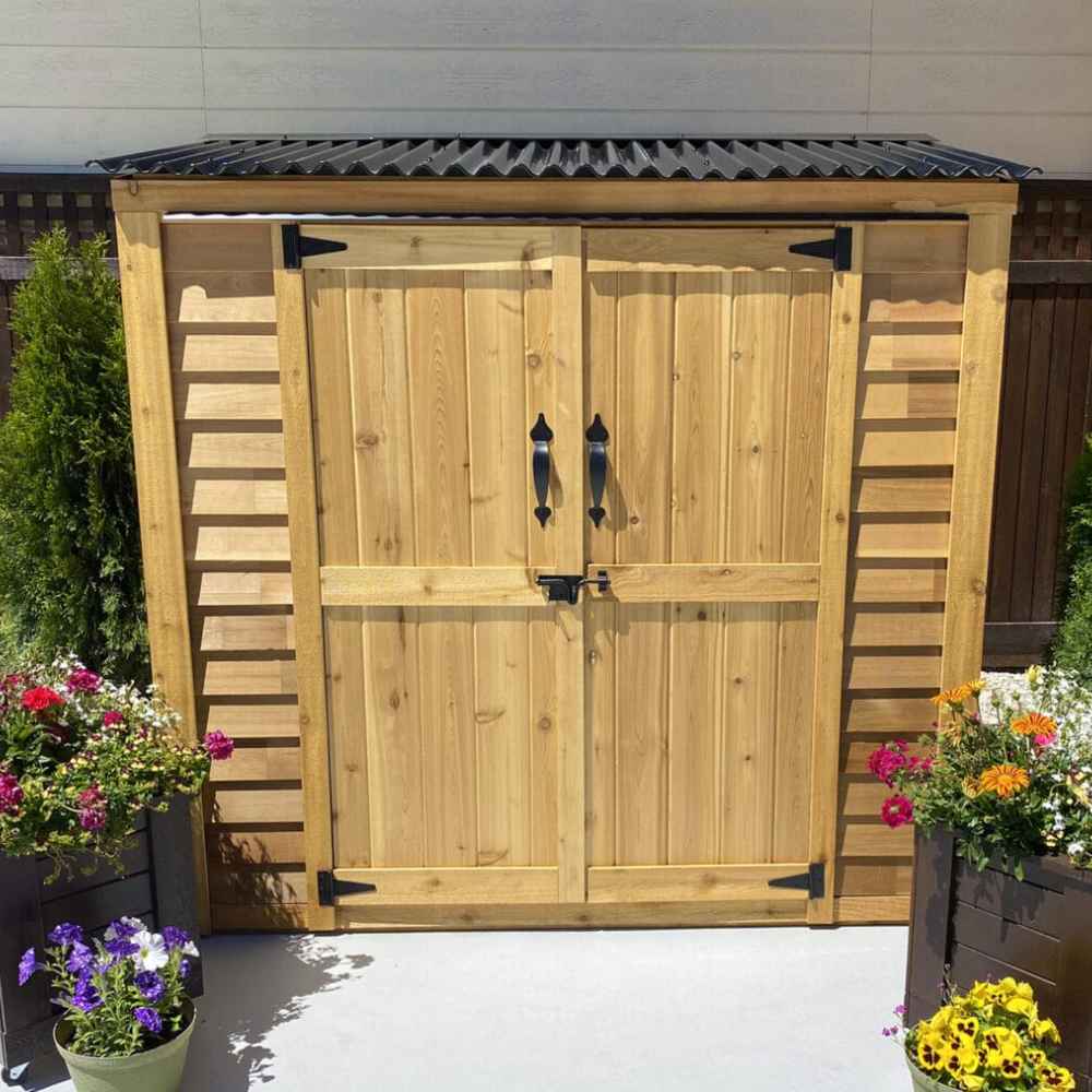 A wooden double-door shed with black metal roof sits surrounded by colorful planters, showcasing the Outdoor Living Today Grand Garden Chalet