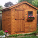 A wooden shed with a dark stain, front door, and window flower box with pink flowers, standing in a garden, showing the Outdoor Living Today 8×8 Gardener Shed