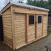 A wooden shed with double sliding doors and two windows stands on a stone path, showcasing the rustic charm of the Outdoor Living Today GardenSaver 12×4 with Sliding Doors