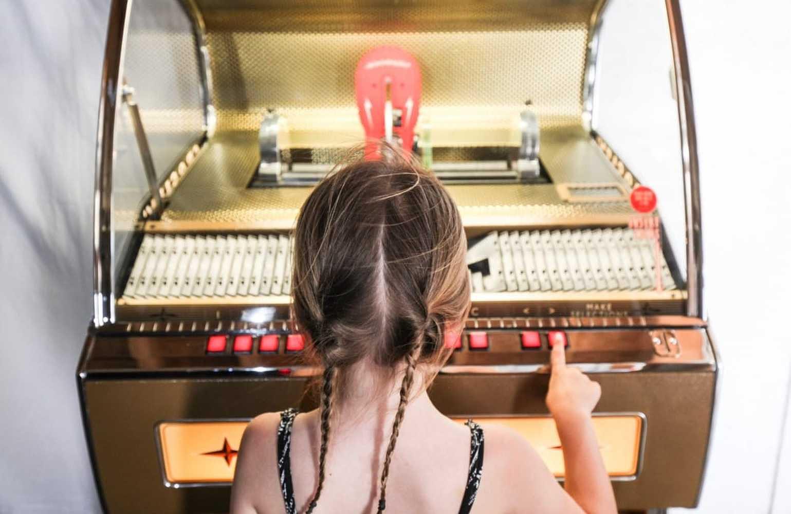 A young girl with braided hair stands in front of the Crosley Rocket 80 CD Bluetooth Full-Size Jukebox - Oak, pressing one of its vibrant red buttons