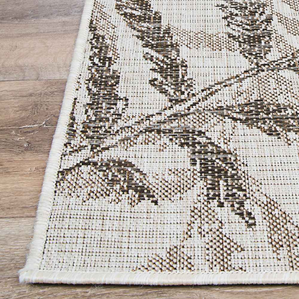A zoomed-in view shows the neat edge stitching and brown-toned leaf design of the Couristan Charm Indoor/Outdoor Rug - Kimberly/Naturals against a wood floor