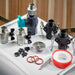 Ball valves, black hose connectors, a stainless drain grate, red O-rings and thread seal tape for Golden Designs Dynamic Cold Therapy Barrel 304 Stainless Steel Cold Plunge rest on a white surface